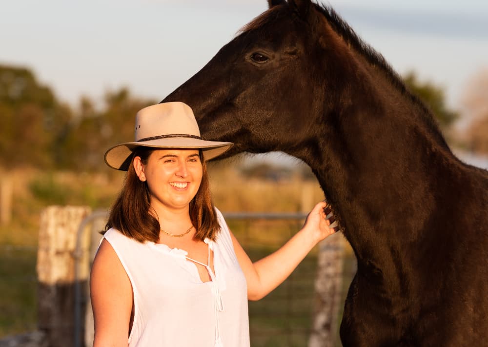 Woman in white dress and Akubra stands with brown horse at sunset.