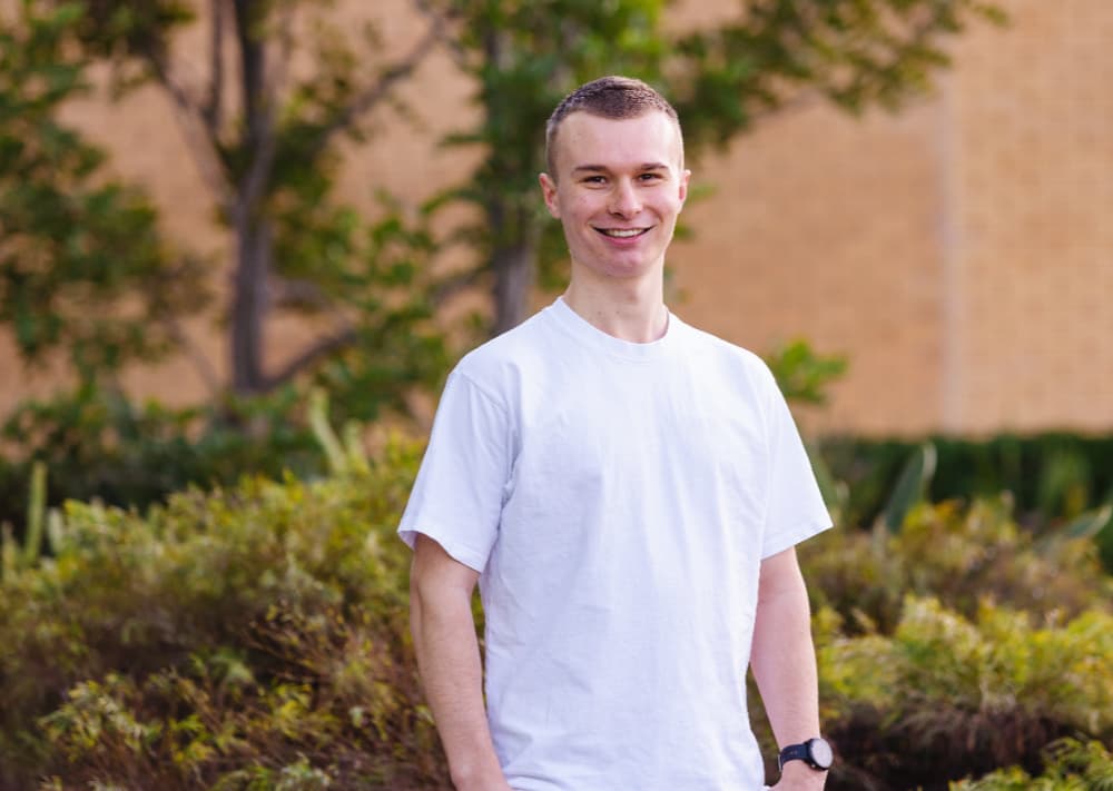 University student stands in front of shrubbery in white shirt