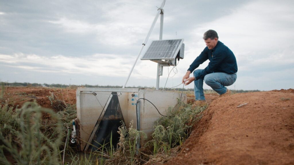 Peter person kneels beside a solar-powered monitoring unit in a grassy field, surrounded by vegetation. The unit includes a metal box with cables and pipes, partially buried in the ground, and a solar panel mounted on a pole above it. The scene illustrates sustainable technology used for environmental or agricultural research.