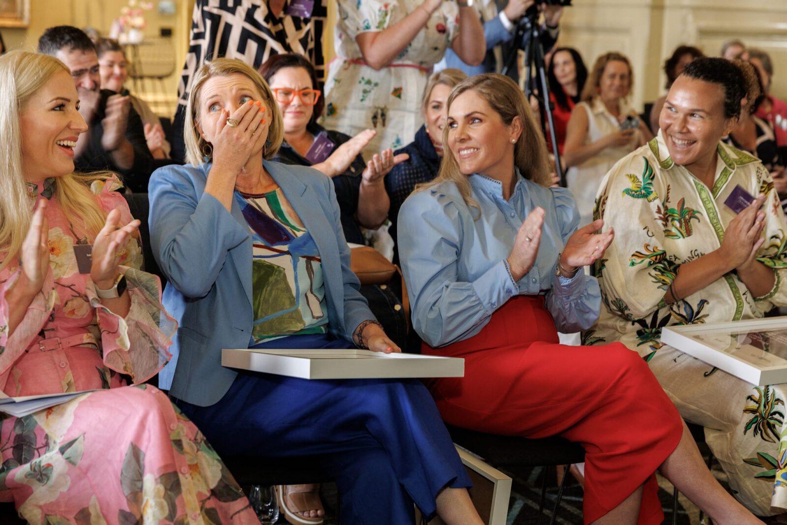 A group of women sit and clap at an indoor agriculture event. One woman covers her mouth in surprise or joy, while the others smile and applaud, creating a celebratory atmosphere with a focus on rural community achievements.