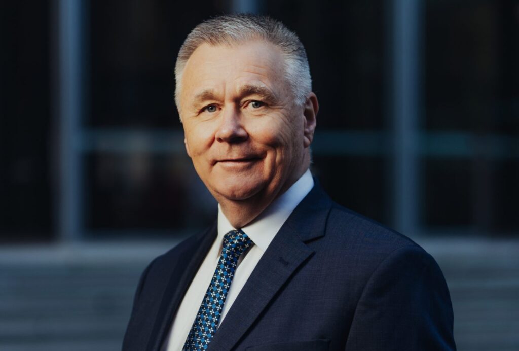 Mick Veitch, AgriFutures Australia Chair. Mick is wearing a dark suit with a white shirt and blue tie against a dark background with a building in the background.