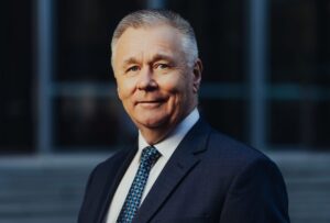 Mick Veitch, AgriFutures Australia Chair. Mick is wearing a dark suit with a white shirt and blue tie against a dark background with a building in the background.