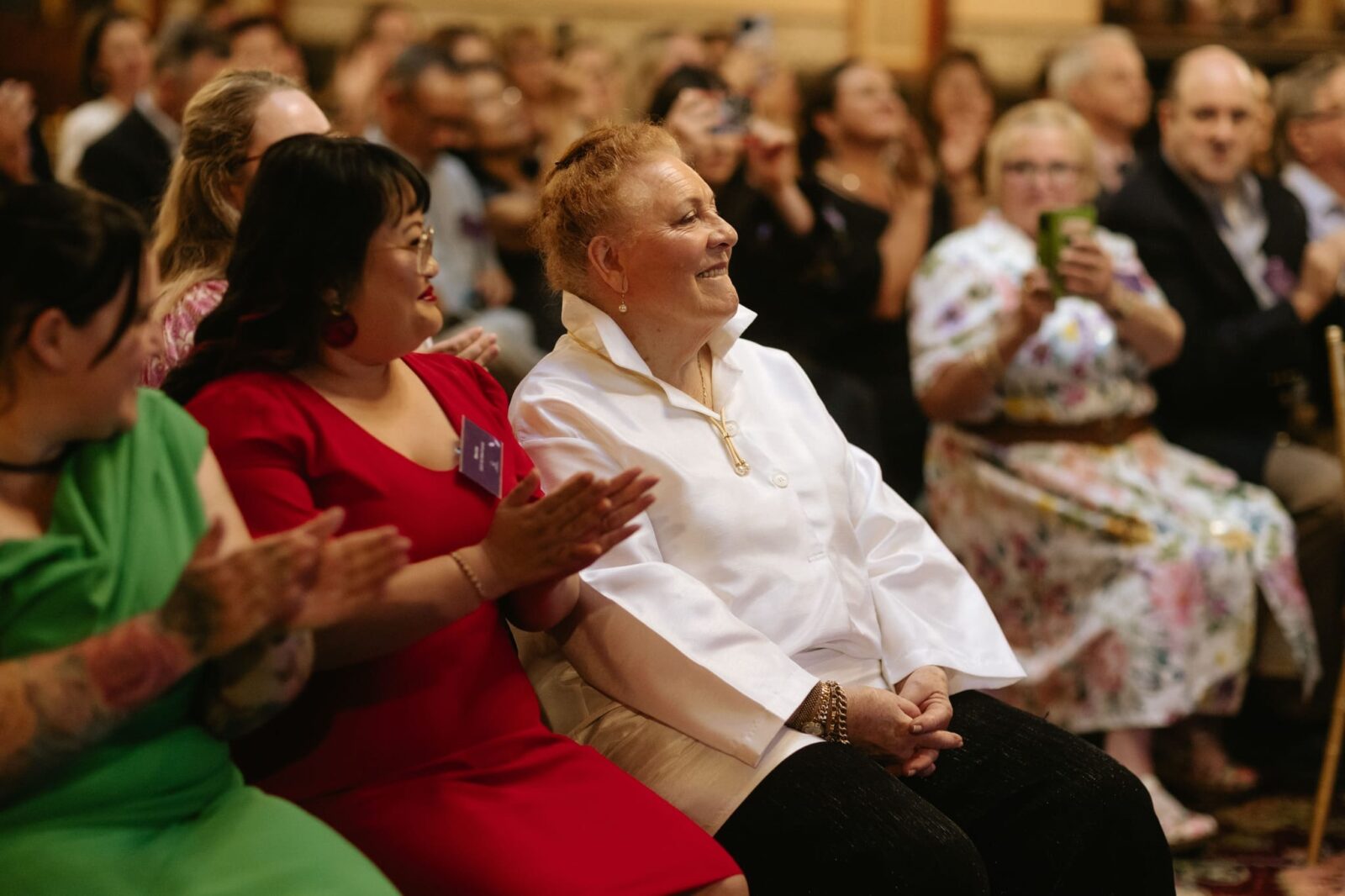 A smiling elderly woman in a white shirt sits among a clapping, diverse crowd at an indoor event. People around her, some from rural backgrounds, are dressed formally and appear engaged and joyful.