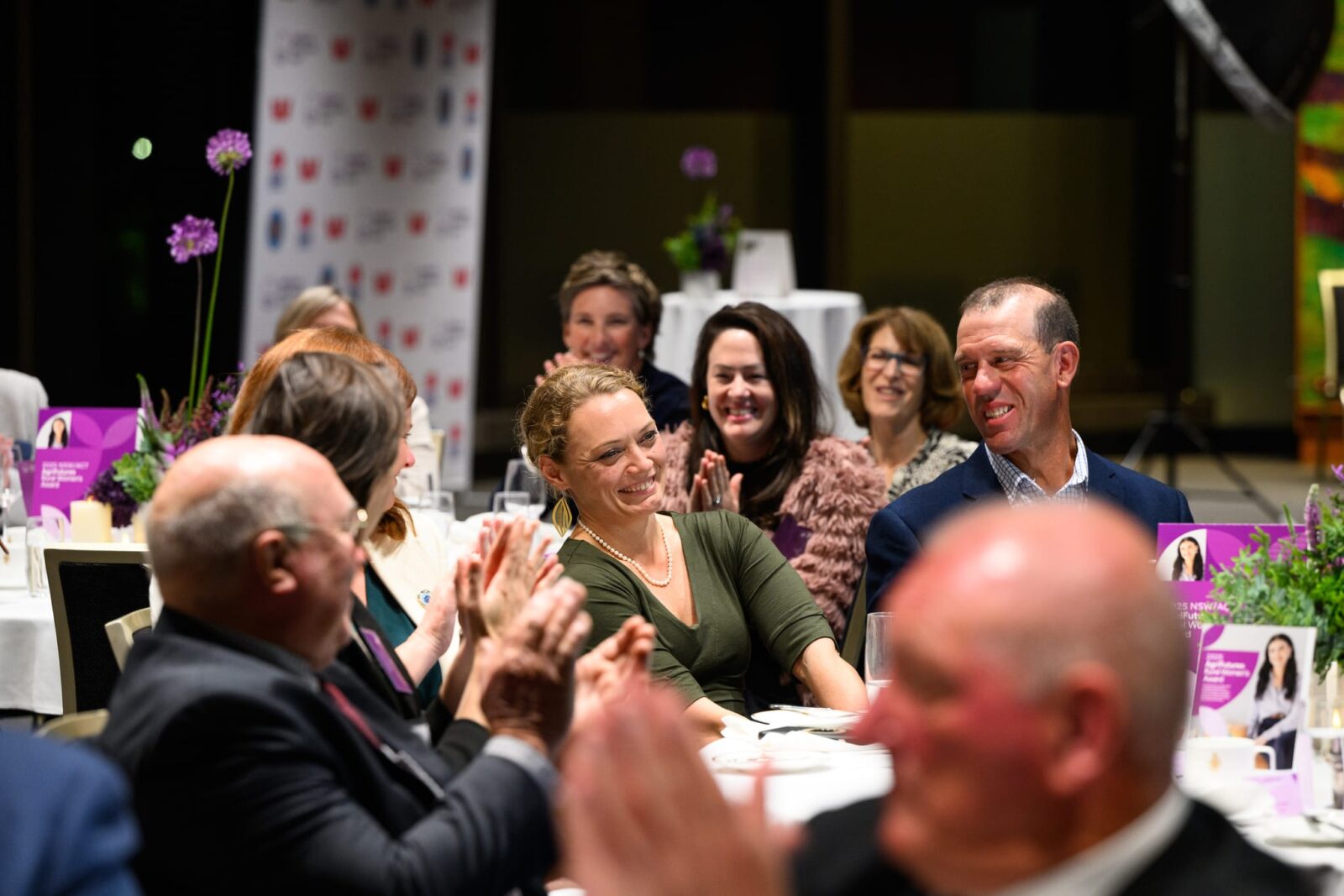 People in formal attire sit at decorated tables, clapping and smiling during an indoor event celebrating development in Australia. Some chat and laugh, with colorful programs and floral centerpieces visible on the tables.