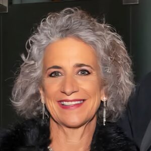 A smiling older woman with curly gray hair, wearing dangly earrings and a black fuzzy top, stands in front of a dark background—her passion for agriculture and research shining through her warm expression.