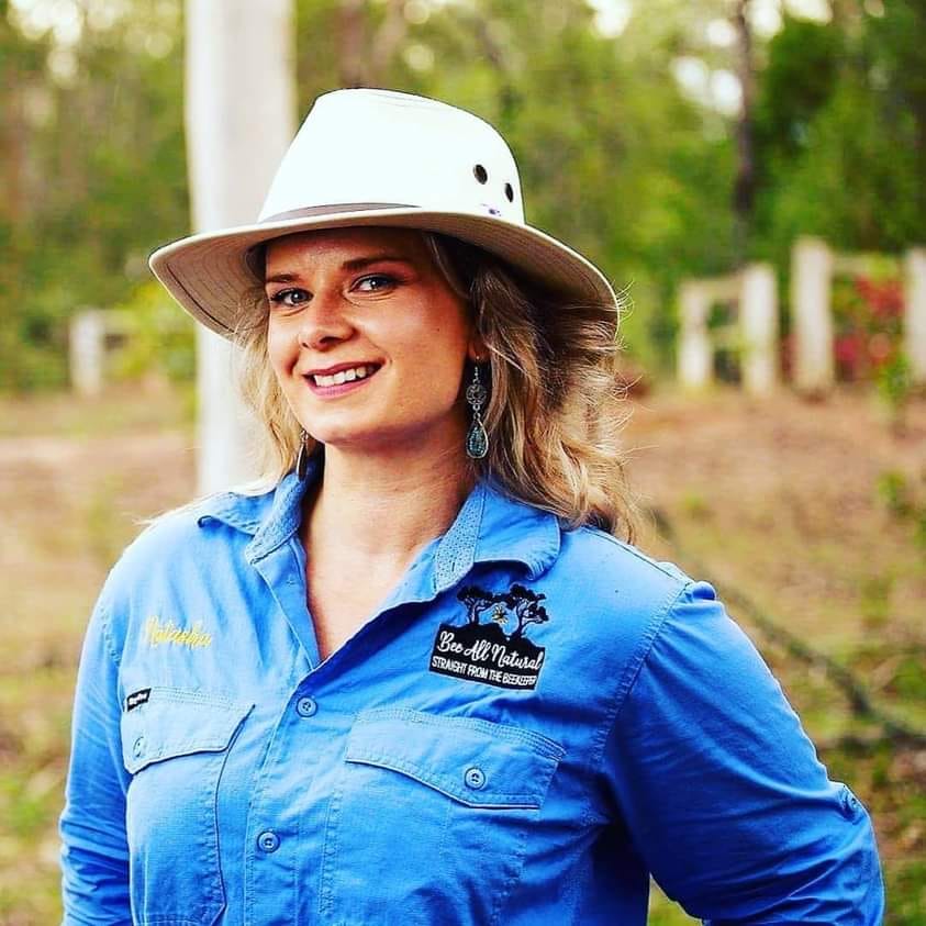 A woman wearing a white wide-brimmed hat and blue button-up shirt stands outdoors in rural Australia, smiling. There is greenery and a wooden fence in the blurred background. Her shirt has embroidered text and a logo on the chest.