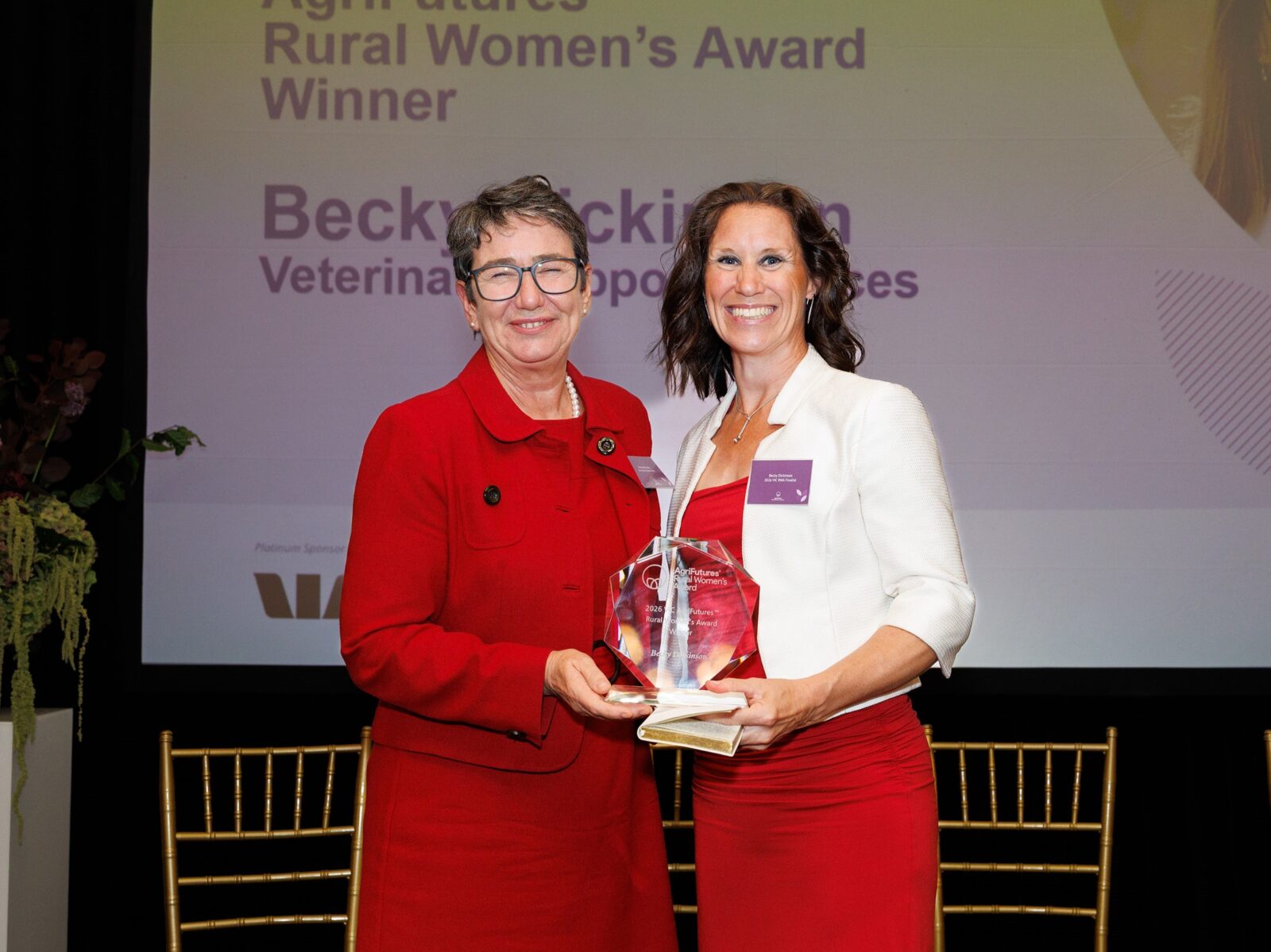 Becky Dickinson stands on stage receiving the Victorian AgriFutures Rural Women’s Award, holding a glass trophy alongside the presenter, with an event screen displaying the Award title behind them.