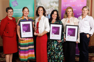 Six women stand together at an indoor awards ceremony, holding framed certificates. They are dressed in formal attire and positioned in front of AgriFutures Rural Women’s Award and Victorian Rural Women’s Network branded backdrops.