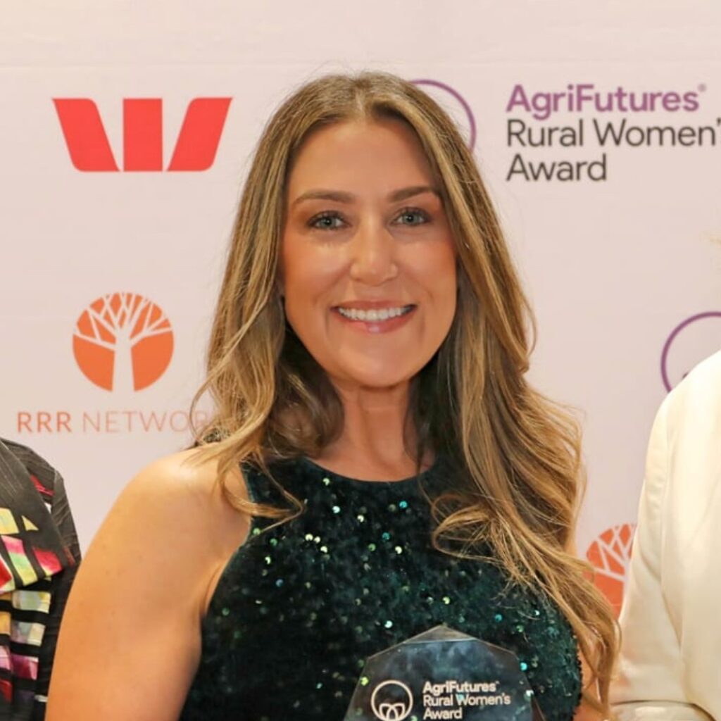 A smiling woman with long brown hair holds an AgriFutures Rural Women’s Award trophy, celebrating excellence in agriculture, standing before a backdrop with sponsor logos including Westpac and RRR Network in Australia.