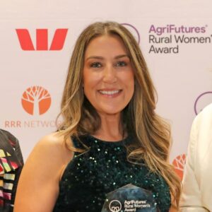 A smiling woman with long brown hair holds an AgriFutures Rural Women’s Award trophy, celebrating excellence in agriculture, standing before a backdrop with sponsor logos including Westpac and RRR Network in Australia.