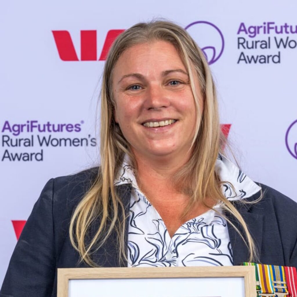 A smiling woman with long blonde hair holds a framed award, celebrating her achievements in agriculture. She is wearing a navy jacket and patterned blouse. The background displays logos for the AgriFutures Rural Womens Award and Westpac.