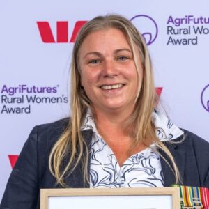 A smiling woman with long blonde hair holds a framed award, celebrating her achievements in agriculture. She is wearing a navy jacket and patterned blouse. The background displays logos for the AgriFutures Rural Womens Award and Westpac.