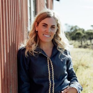 A woman with wavy blonde hair, wearing a dark button-up shirt, stands outside leaning against a red corrugated wall. She is smiling, with a sunny rural landscape and trees in the background.