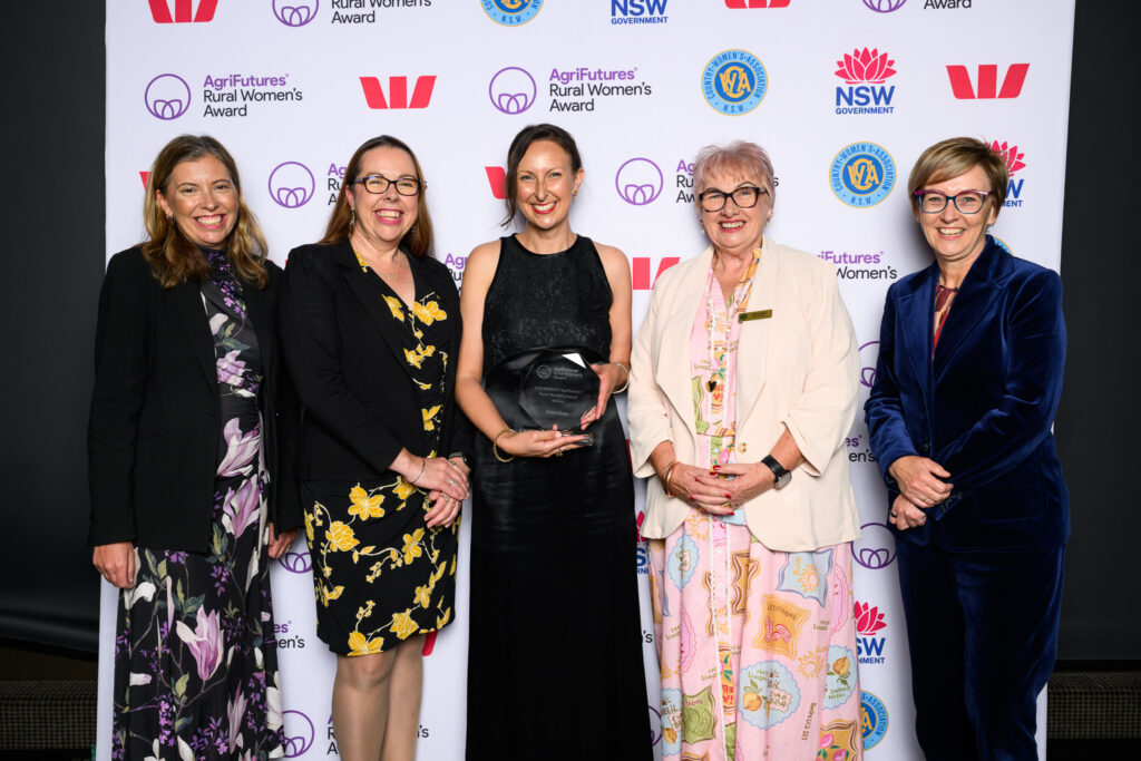 Five women stand together smiling in front of a backdrop featuring logos for the AgriFutures Rural Women’s Award and sponsors. The woman in the centre holds a black award plaque. They are dressed in formal attire.
