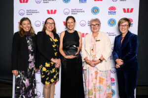 Five women stand together smiling in front of a backdrop featuring logos for the AgriFutures Rural Women’s Award and sponsors. The woman in the centre holds a black award plaque. They are dressed in formal attire.
