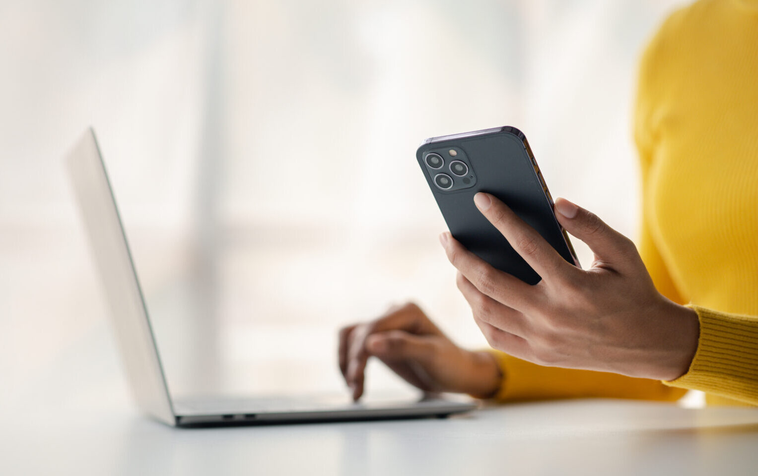 A person in a yellow jumper uses a laptop with one hand and holds a mobile phone in the other, sitting at a white desk—perhaps managing tasks for an SEO agency, with a blurred background.