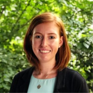 A woman with straight, shoulder-length auburn hair smiles at the camera. She wears a black blazer, a light blue top, and a heart-shaped necklace, standing in front of green, leafy trees.