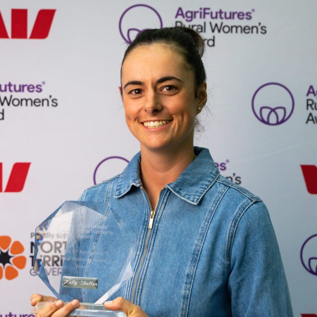 A woman in a blue denim jacket smiles whilst holding an award trophy. She stands in front of a backdrop with logos, including AgriFutures Rural Women's Award and Westpac.
