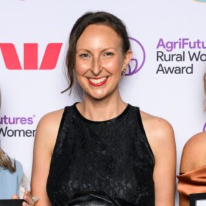 A smiling woman in a sleeveless black dress stands in front of a backdrop with AgriFutures Rural Women’s Award and sponsor logos. She is holding an award and posing for a photo.