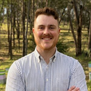A person with short curly hair and a beard, wearing a white and blue striped button-up shirt, stands smiling with arms crossed in a sunlit forest with trees and green grass in the background.