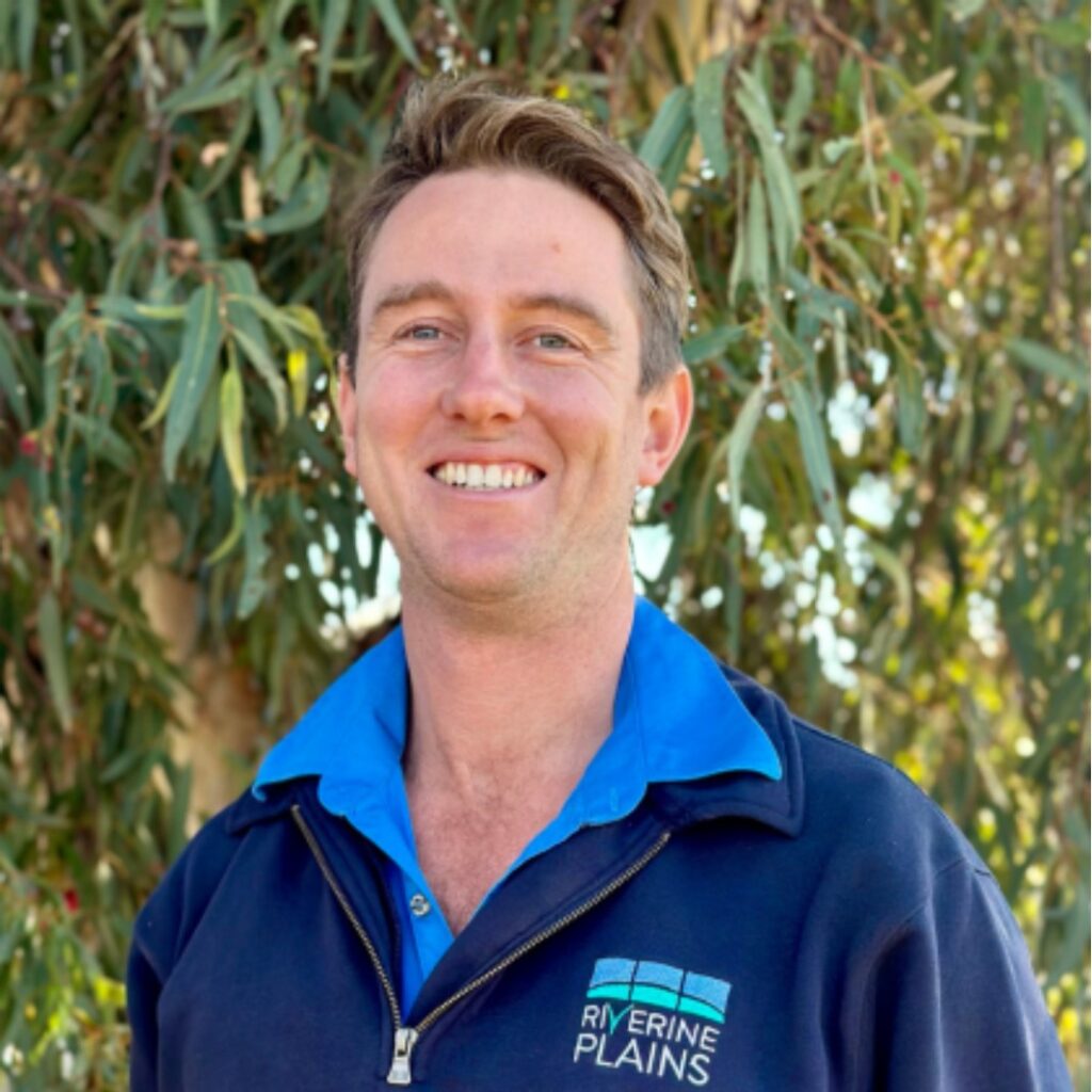 A smiling person wearing a blue collared shirt and a Riverine Plains jacket stands outdoors in front of leafy green trees.