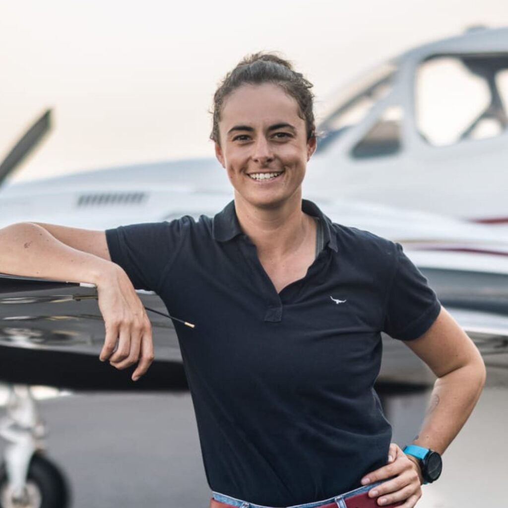 A woman with curly hair tied back is smiling and leaning on the wing of a small aeroplane. She is wearing a navy polo shirt and a watch, and the background shows a blurred aircraft.