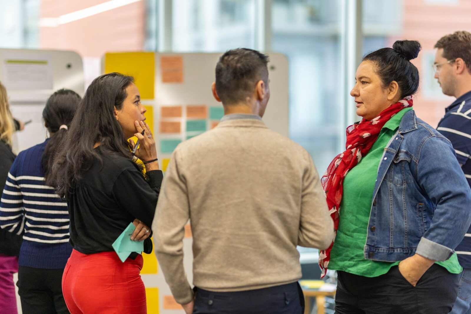 Three people stand in a brightly lit office, engaged in conversation near whiteboards covered with colourful sticky notes. Others are visible in the background, working and talking.