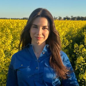 A woman with long brown hair wearing a blue shirt stands in a field of yellow flowers, smiling softly at the camera under a clear sky.