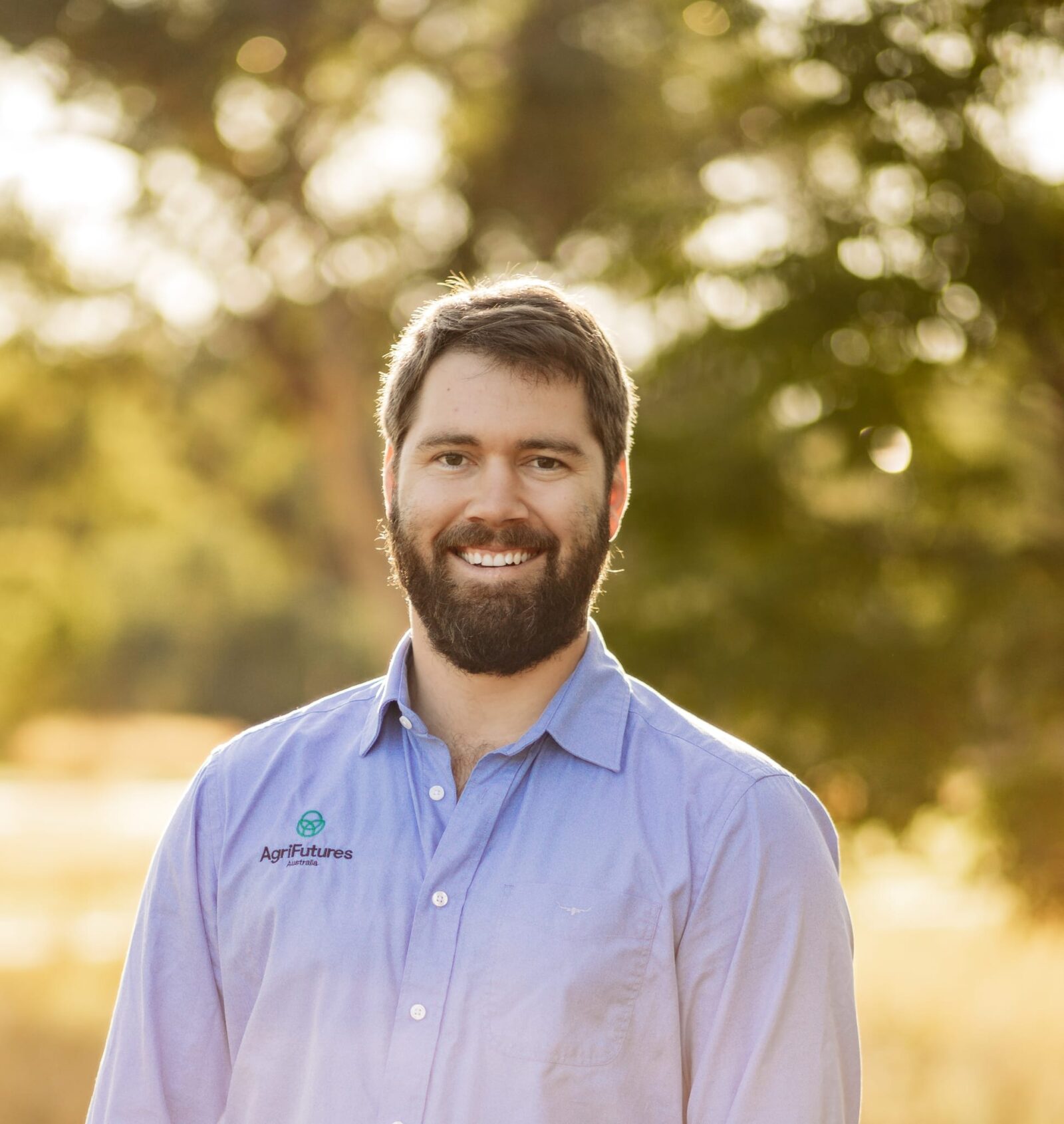 A bearded man with brown hair, wearing a light blue shirt with an AgriFutures logo, stands outdoors smiling, with blurred green trees and sunlight in the background.