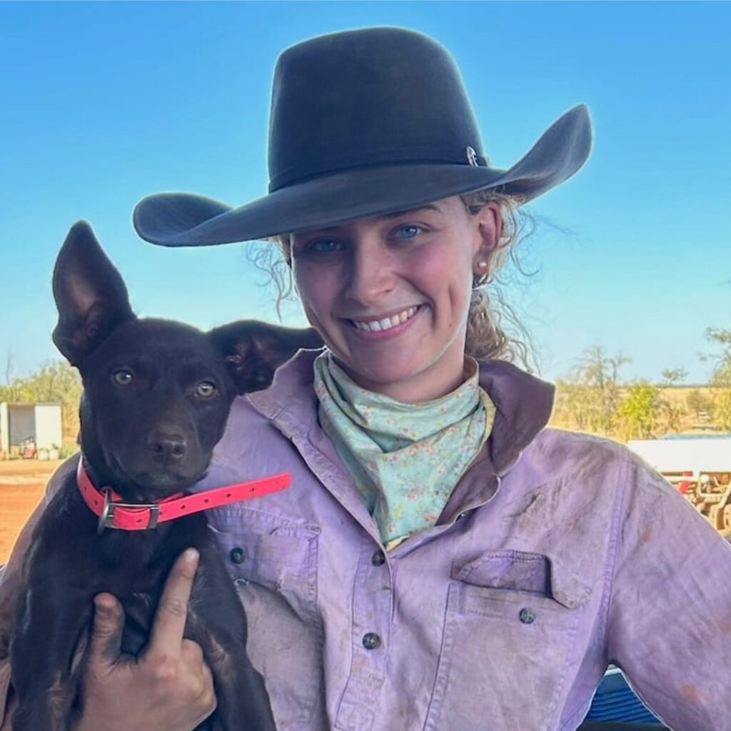 A smiling woman in a cowboy hat and dusty shirt holds a brown dog with large ears and a red collar, standing outdoors under a clear blue sky.