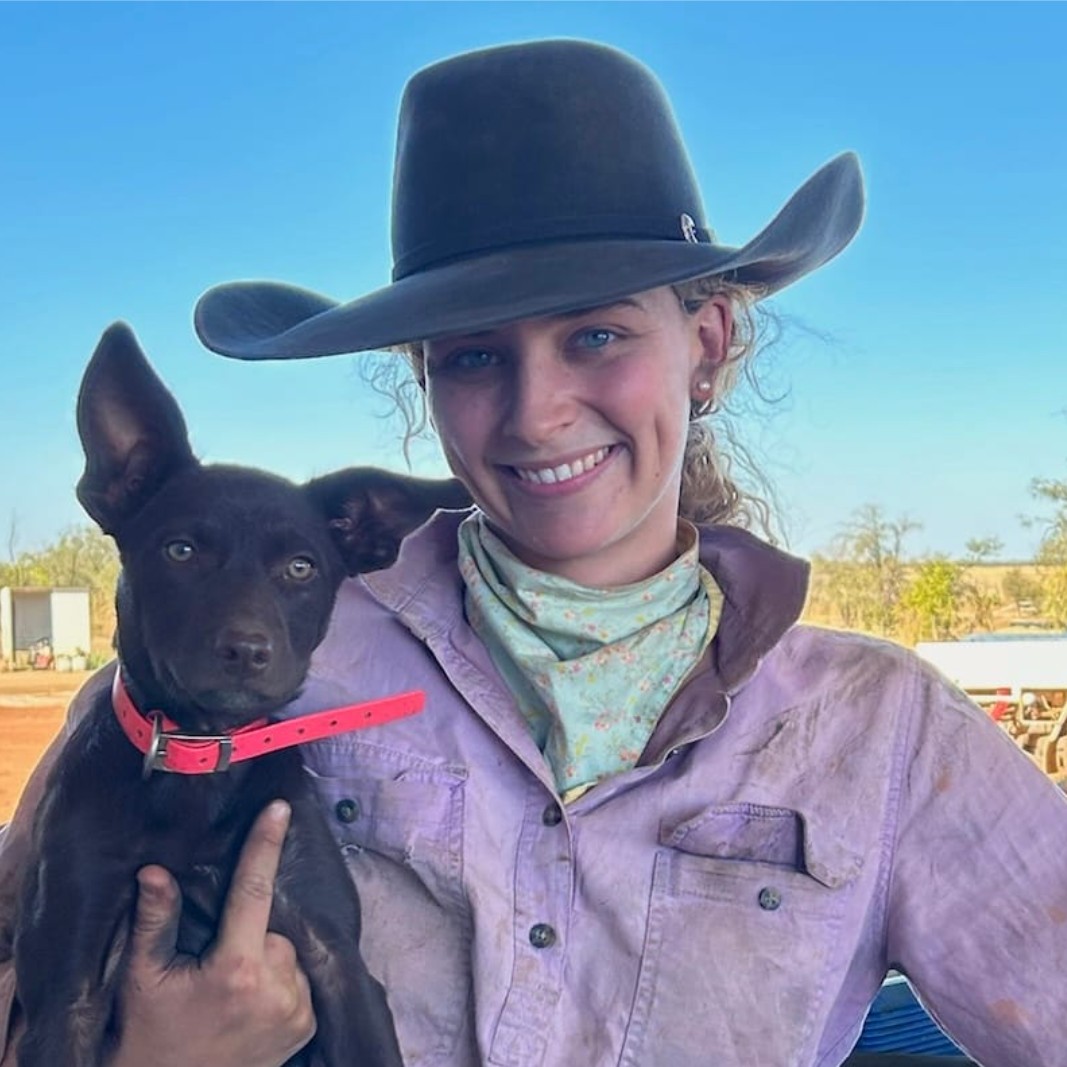 A smiling woman in a cowboy hat and dusty shirt holds a brown dog with large ears and a red collar, standing outdoors under a clear blue sky.