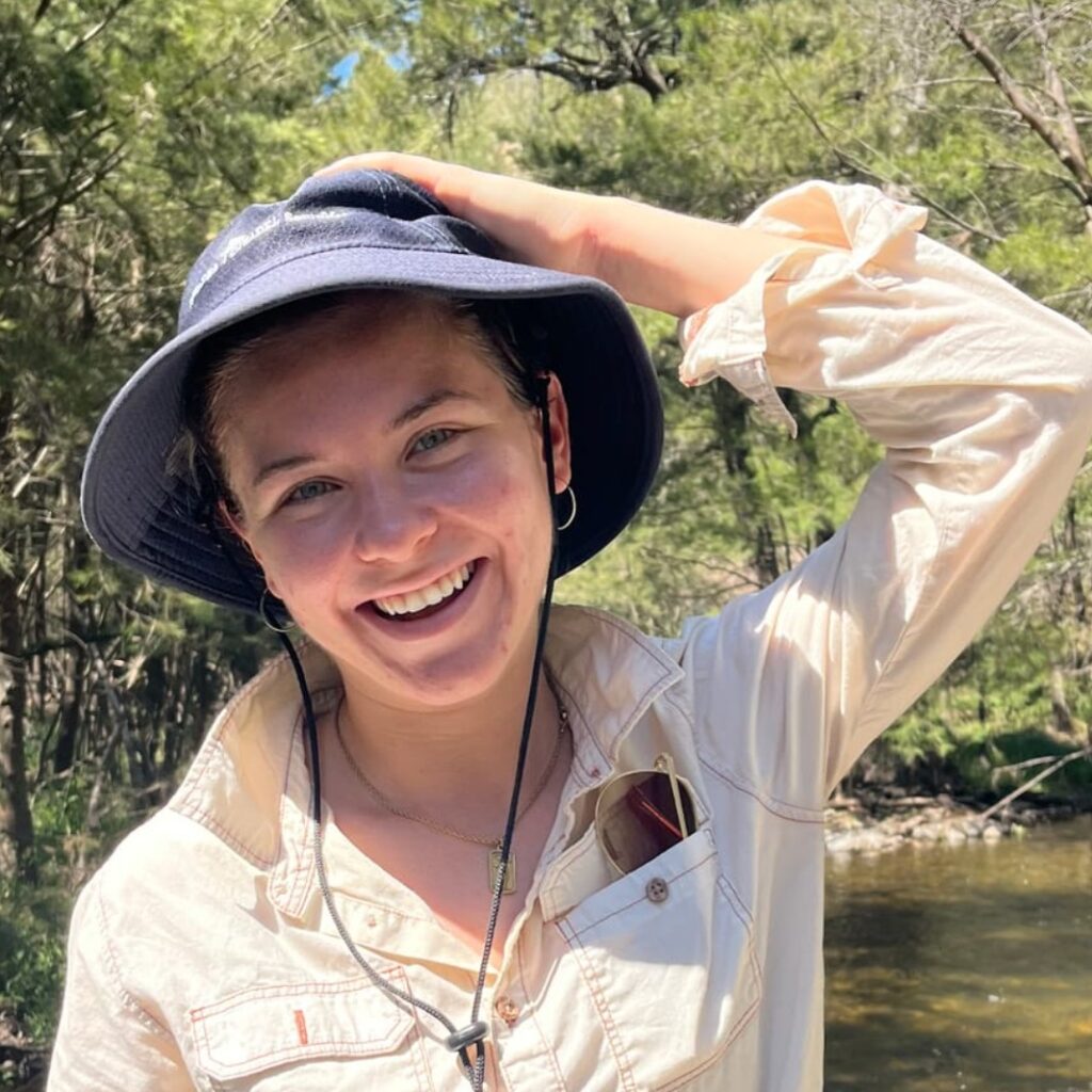 A smiling person wearing a wide-brimmed hat and light-coloured shirt stands outdoors near a flowing stream, with one hand on their hat. Lush green trees and sunlight fill the background.