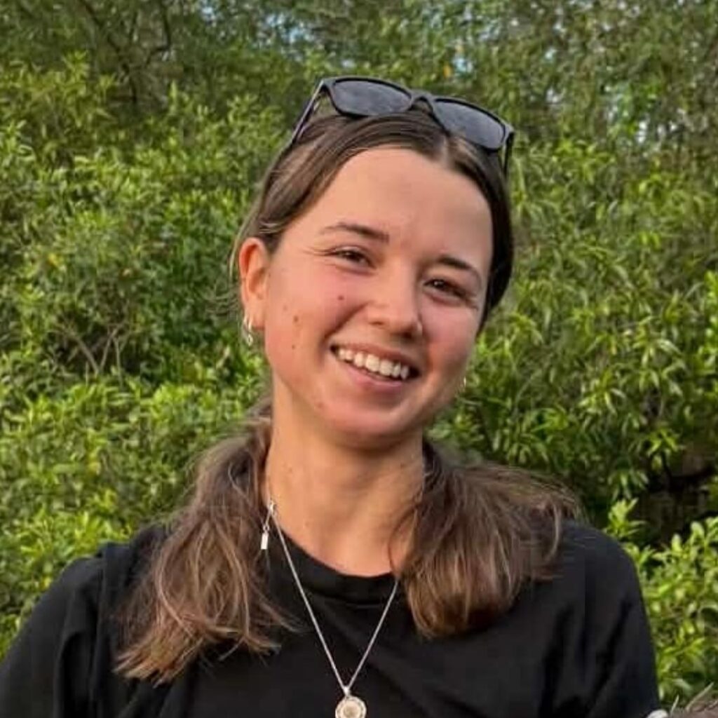 A young woman with long brown hair in plaits, wearing sunglasses on her head, a black shirt, and necklaces, smiles outdoors with green trees in the background.