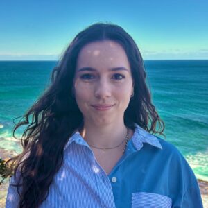 A young woman with long dark hair stands outdoors, smiling softly, with the sea and blue sky in the background. She is wearing a blue and white button-up shirt and a gold necklace.