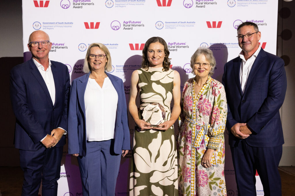 Five people stand posing for a photo at an awards event. The woman in the centre holds a glass trophy, celebrating her RWA achievement. A branded backdrop with logos for the AgriFutures Rural Women’s Award is behind them.
