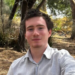 A young man with short brown hair and light facial hair, wearing a light grey shirt, sits outdoors in a wooded area with sunlight filtering through the trees.