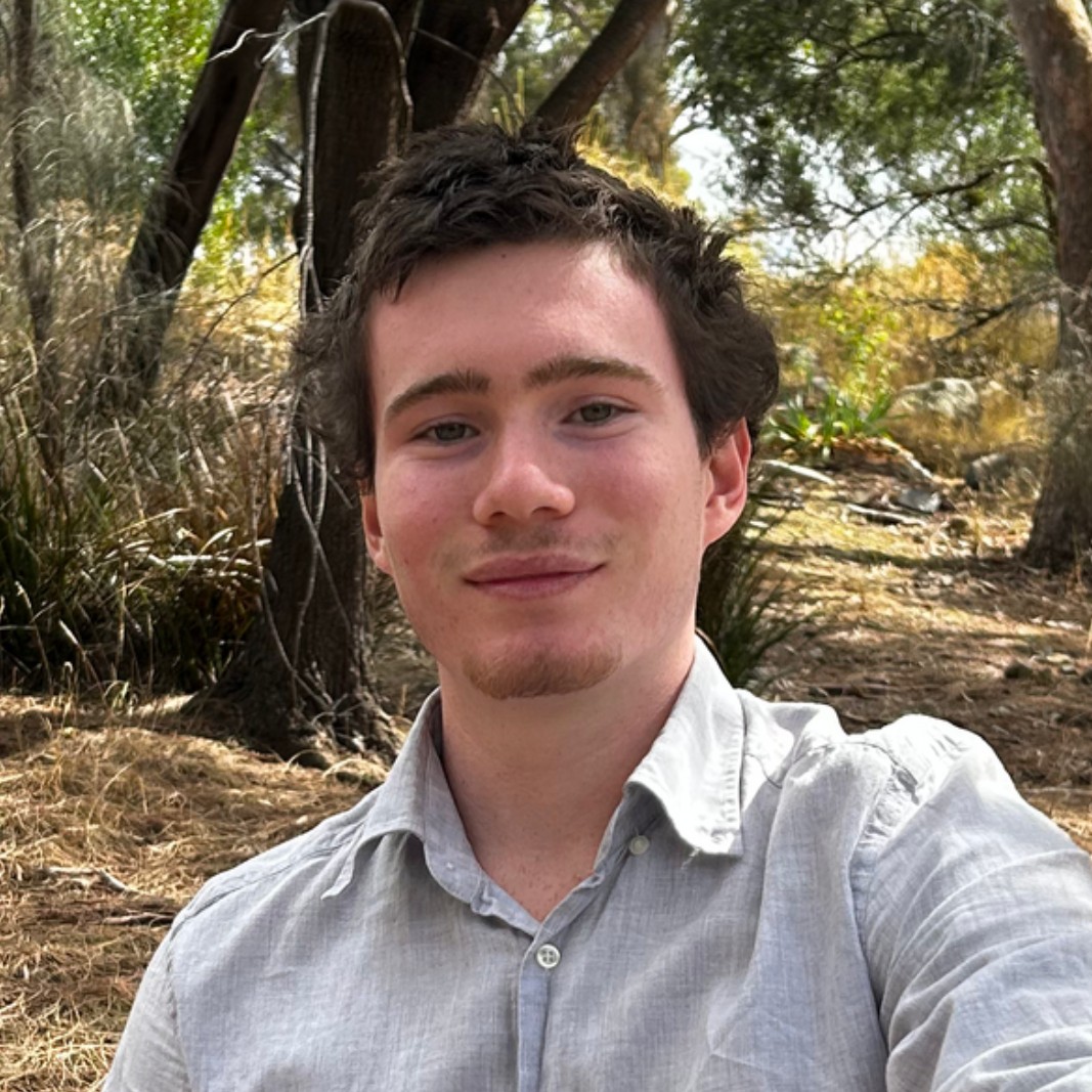 A young man with short brown hair and light facial hair, wearing a light grey shirt, sits outdoors in a wooded area with sunlight filtering through the trees.