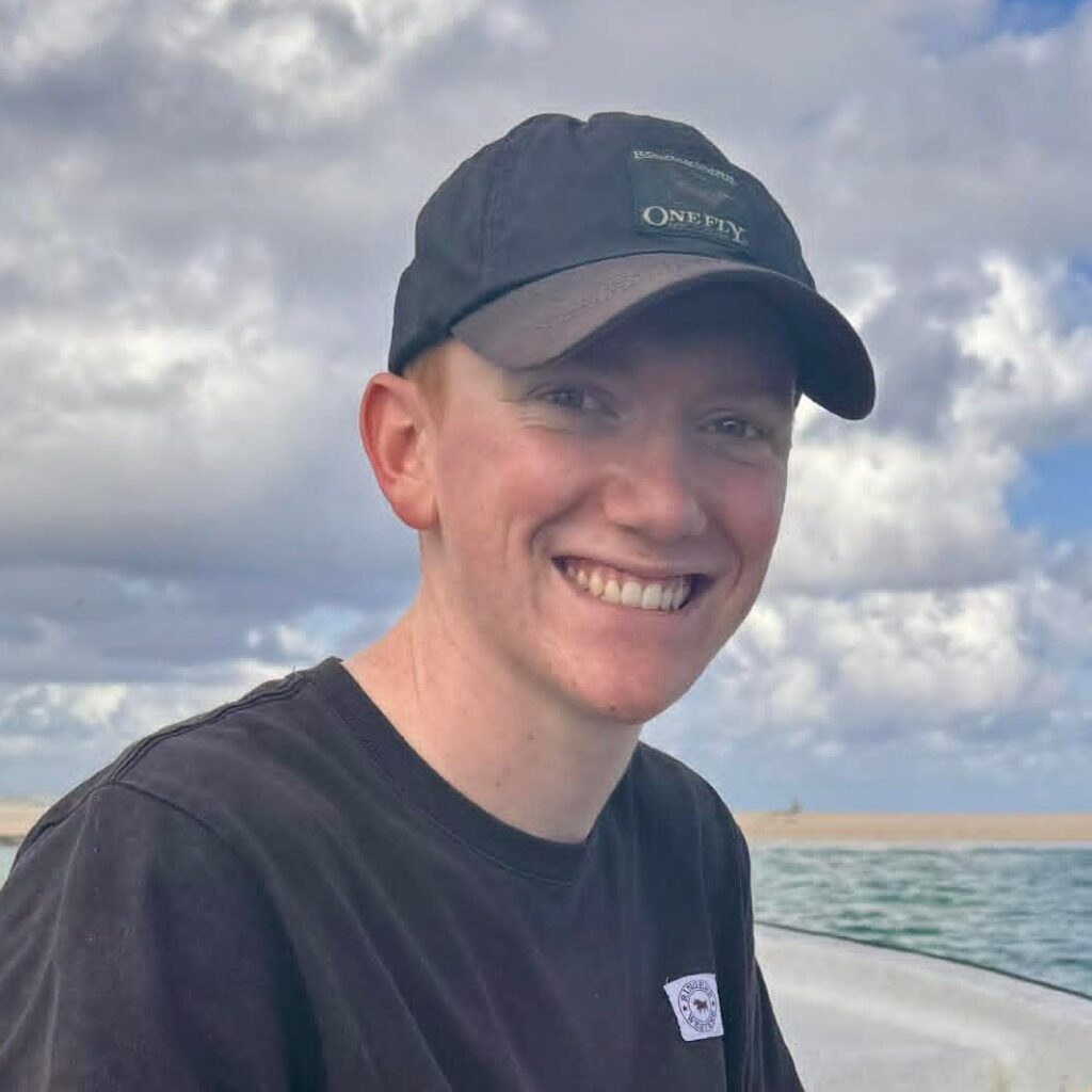 A smiling person wearing a black cap and T-shirt sits outdoors on a boat with water and a cloudy sky in the background.