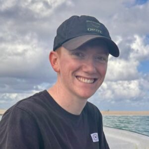 A smiling person wearing a black cap and T-shirt sits outdoors on a boat with water and a cloudy sky in the background.