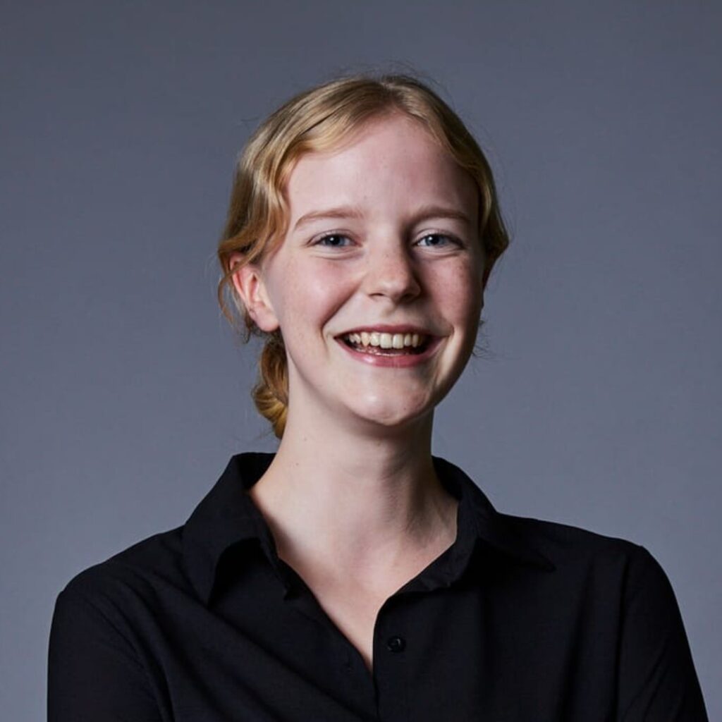 A young woman with blonde hair tied back, wearing a black collared shirt, smiles warmly against a plain grey background.