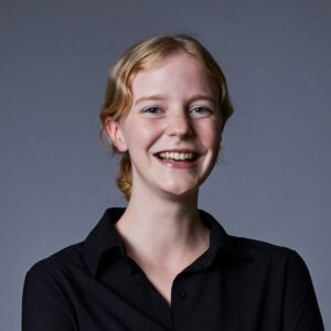 A young woman with blonde hair tied back, wearing a black collared shirt, smiles warmly against a plain grey background.