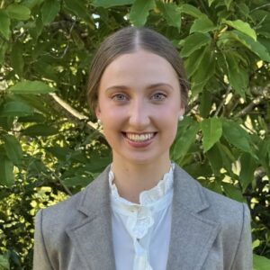 A young woman with light brown hair tied back, wearing a grey blazer and white blouse, smiles at the camera whilst standing outdoors in front of leafy green trees.