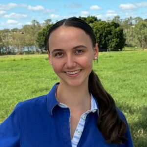 A young woman with dark hair in a low ponytail smiles outdoors, wearing a blue collared shirt. She stands in a grassy field with trees and a blue sky with scattered clouds in the background.