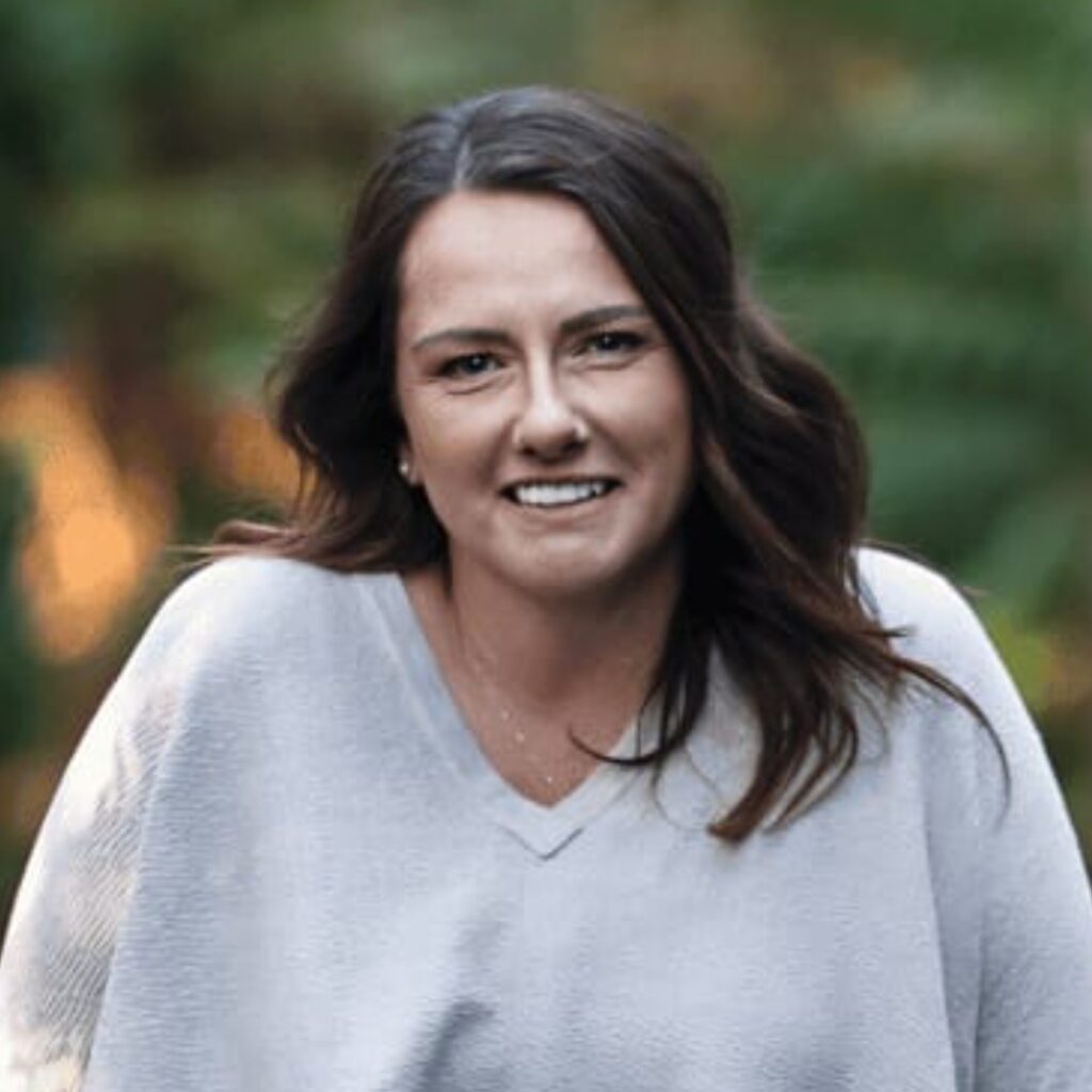 A woman with long brown hair wearing a light grey jumper smiles outdoors with green foliage blurred in the background.