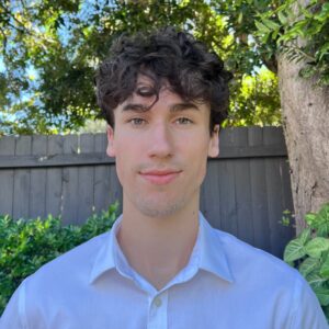 A young man with curly brown hair wearing a white collared shirt stands outdoors in front of a wooden fence, green shrubs, and trees on a sunny day.