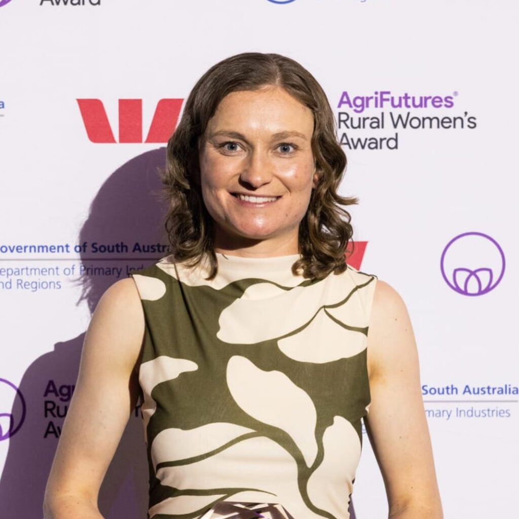 A woman with shoulder-length brown hair smiles at the camera, standing in front of a backdrop with logos including “AgriFutures Rural Women’s Award” and “Westpac.” She wears a sleeveless patterned dress.