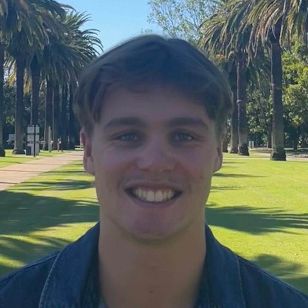 A young man with short brown hair smiles at the camera. He is standing outdoors on a sunny day, with palm trees and a path visible in the background.