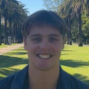 A young man with short brown hair smiles at the camera. He is standing outdoors on a sunny day, with palm trees and a path visible in the background.