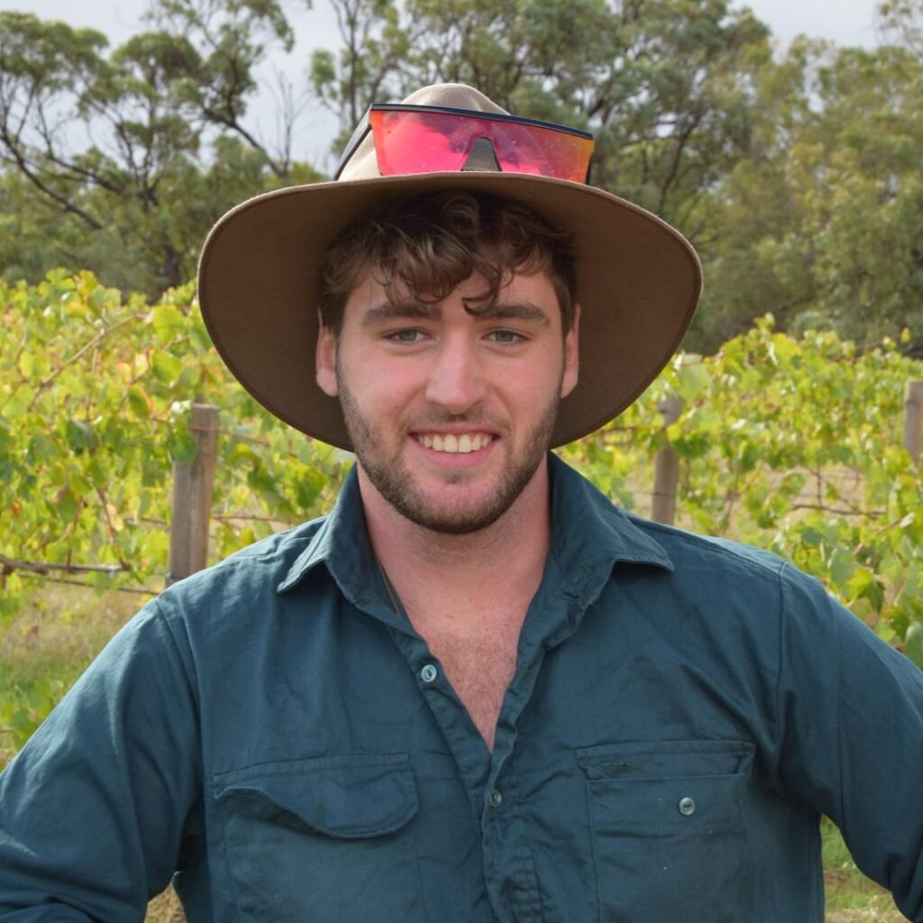 A young man with a beard and tousled hair, wearing a wide-brimmed hat with sunglasses on top and a teal shirt, stands outdoors in front of grapevines and trees, smiling at the camera.