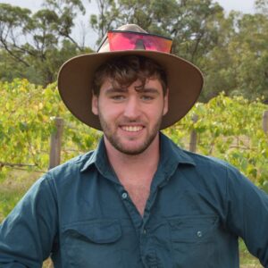 A young man with a beard and tousled hair, wearing a wide-brimmed hat with sunglasses on top and a teal shirt, stands outdoors in front of grapevines and trees, smiling at the camera.